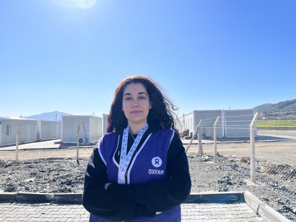 Seda Karakuş in front of a container city wearing a blue vest with OXFAM logo