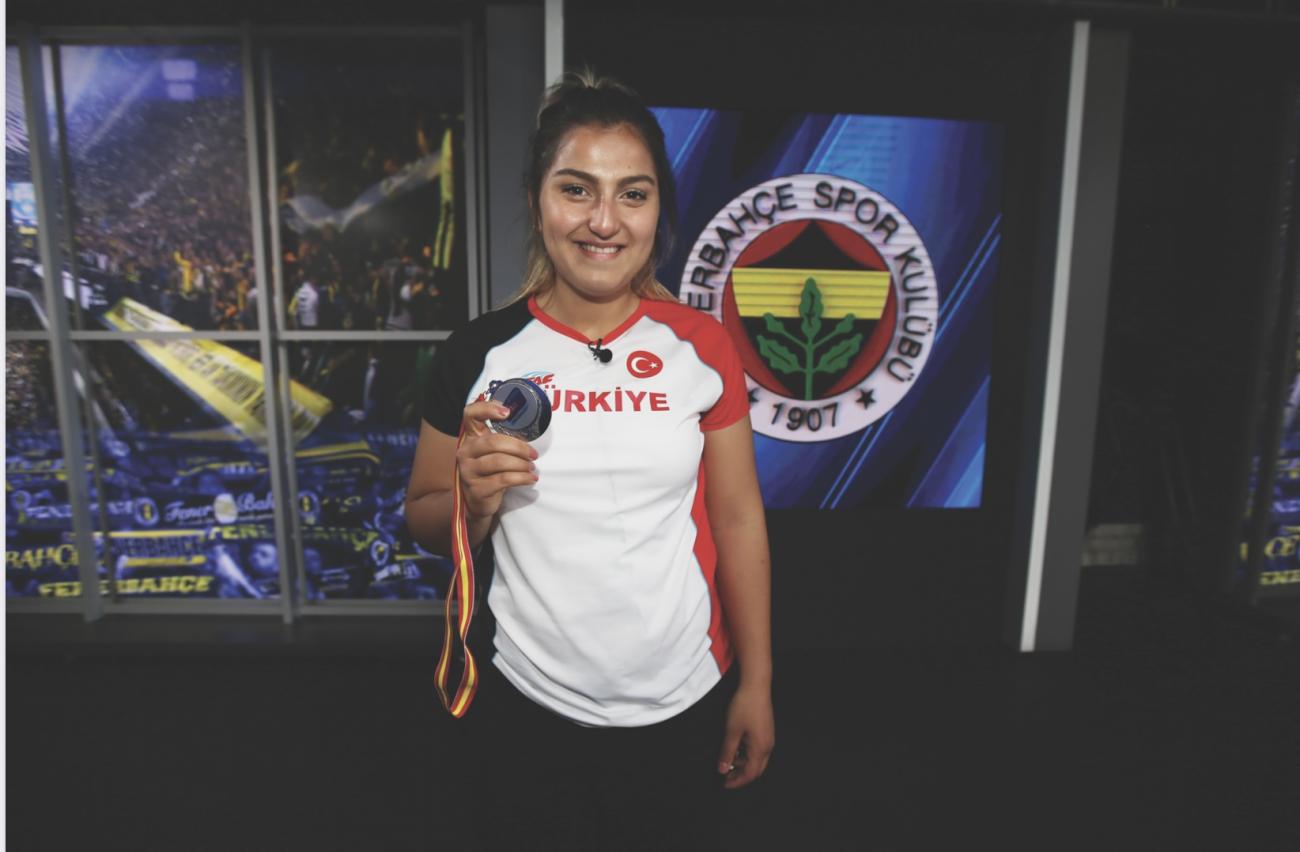 Üzerinde Türkiye yazan t-shirtü ile Eda Tuğsuz Fenerbahçe Spor Kulübü arması önünde madalyasını göstererek poz vermiş 