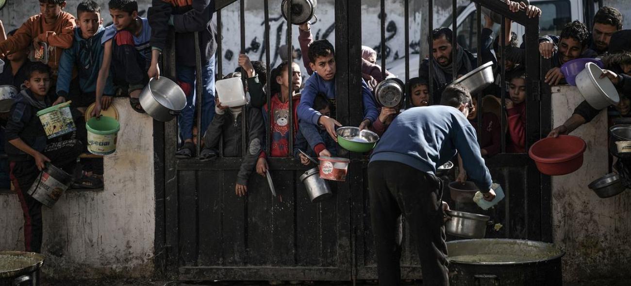 Demir kapının arkasından ve duvarların üzerinden çocuklar yemek dağıtan görevliye doğru yemek kaplarını uzatıyorlar.