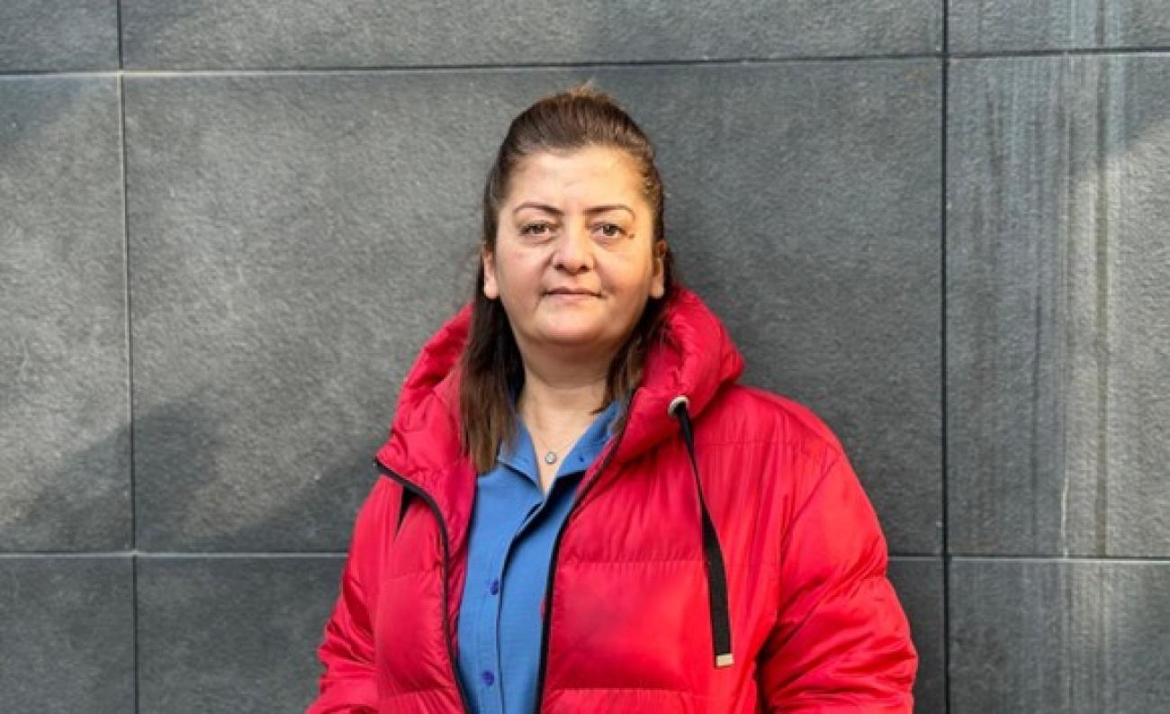 Nesrin Deli mavi gömlek ve kırmızı palto giymiş ve gri bir duvarın öünde ayakta duruyor in blue shirt and red coat standing in front of a dark grey wall