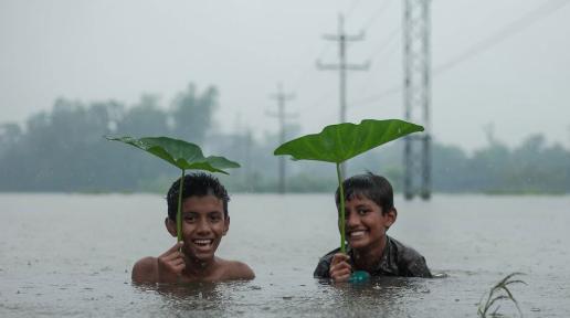 2 children sokaed in water are holding big green leaves on top of their heads under heavy rain 