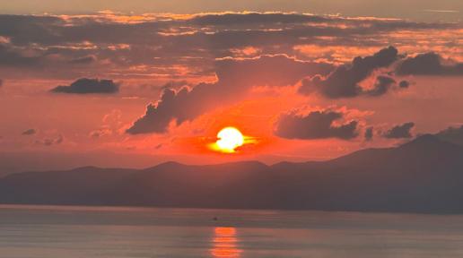 Sun sets behind clouds, hills withs its reclection on the sea