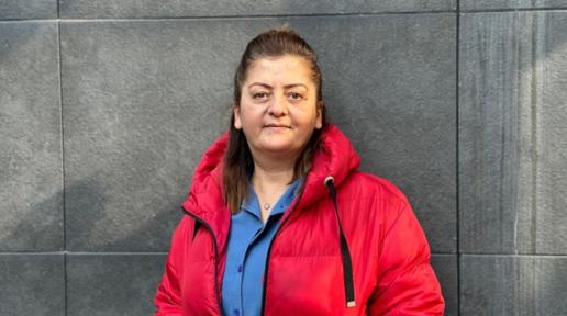 Nesrin Deli mavi gömlek ve kırmızı palto giymiş ve gri bir duvarın öünde ayakta duruyor in blue shirt and red coat standing in front of a dark grey wall